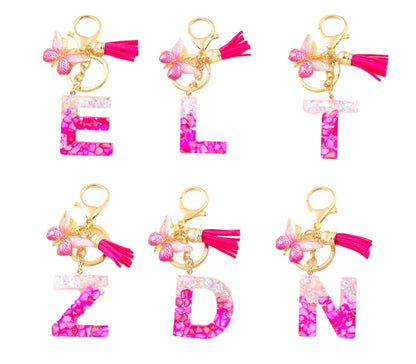 Pink resin alphabet keychains with gold rings, pink tassels, and butterfly charms in letters E, L, T, Z, D, and N