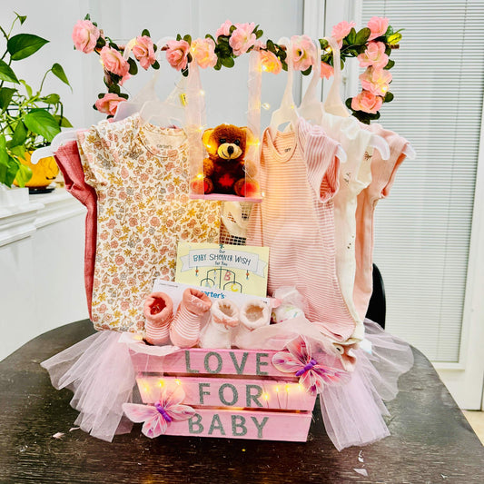 Custom baby shower gift basket with handmade newborn essentials including onesies, socks, booties, plush toy, and wishing card in a decorative pink crate with floral accents.