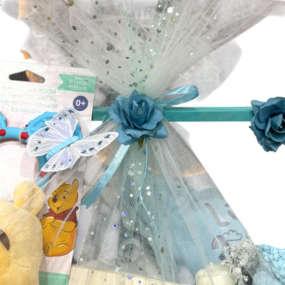 Customizable gift basket for mom with baby clothes, blanket, shea soap, hand-poured candle, and floral bouquet wrapped in decorative blue and white tulle with blue flowers.