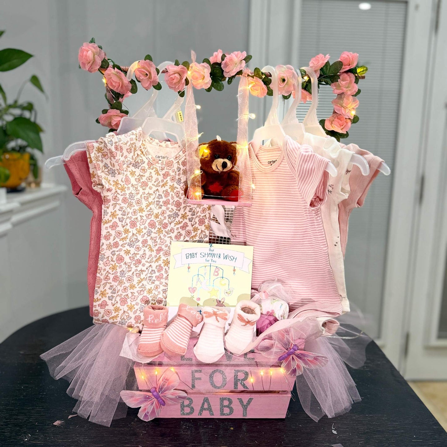 Custom baby shower gift basket with handmade newborn essentials including onesies, booties, socks, plush toy, and wishing card, decorated with pink flowers and tulle.