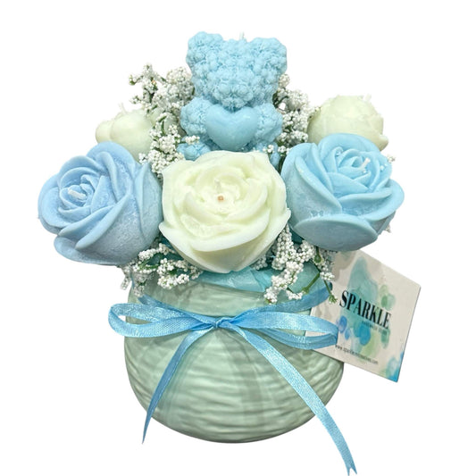 Handmade candle flower bouquet with blue and cream rose-shaped candles, baby’s breath, and a blue teddy bear candle in a decorative round vase with blue ribbon.