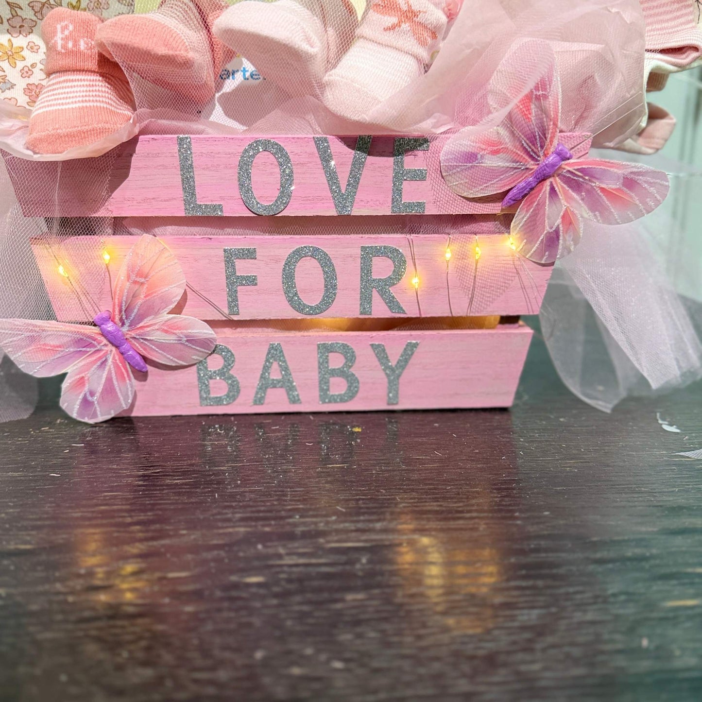Pink wooden crate with "Love for Baby" text, decorated with butterflies, fairy lights, and baby socks, part of a custom baby shower gift basket.