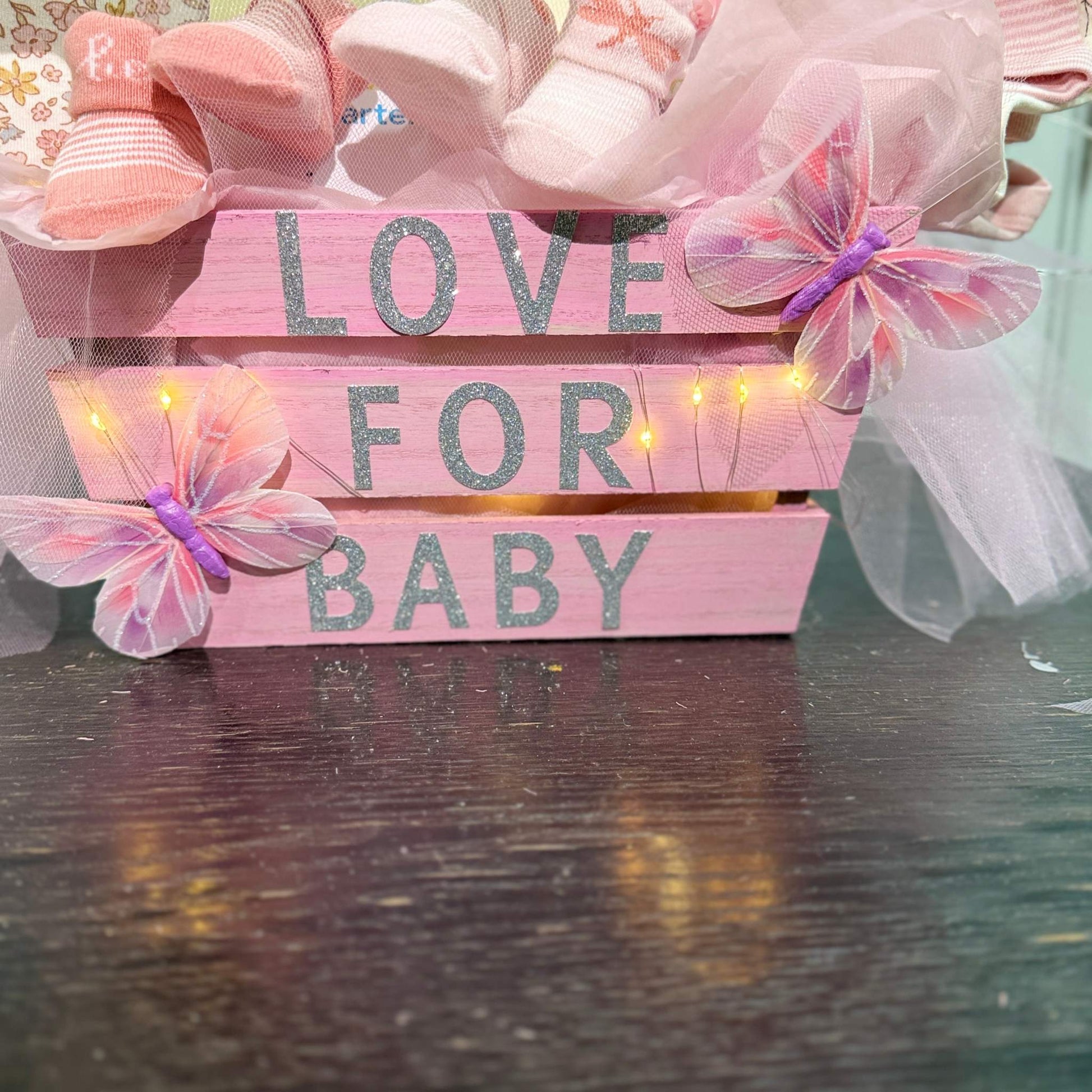 Pink wooden crate with "Love for Baby" text, decorated with butterflies, fairy lights, and baby socks, part of a custom baby shower gift basket.