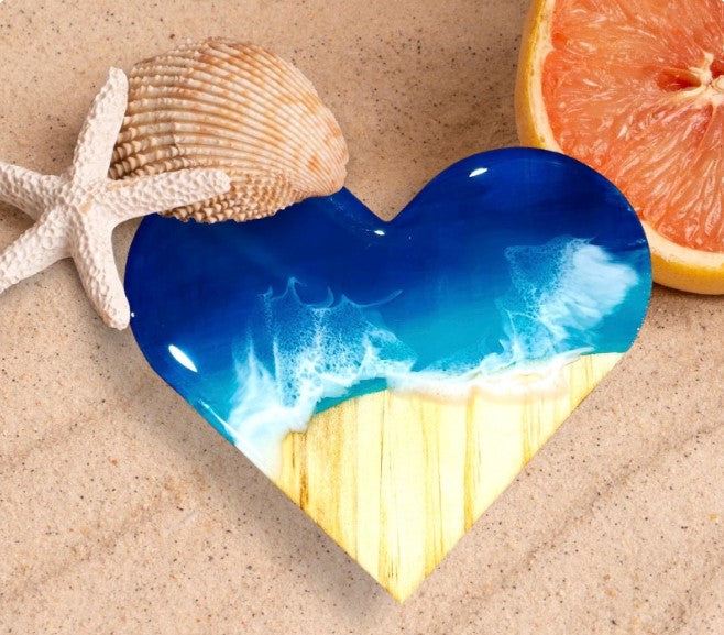 Heart-shaped ocean wave resin coaster with glossy finish, placed on sand with seashell and grapefruit slice nearby.