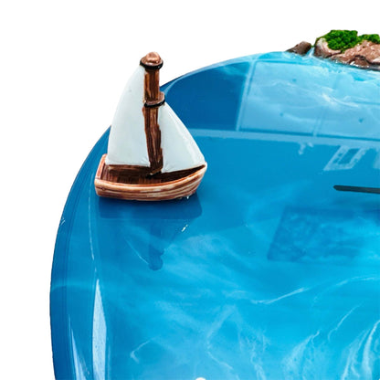 Close-up of unique 3D island wall clock featuring handcrafted clay sailboat on glossy blue resin water surface with part of island and moss visible